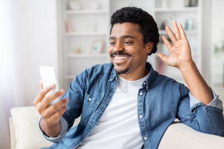 Téléchargez les photos : Heureux afro-américain guy engage dans un appel vidéo de la maison, agitant et souriant, montrant la connectivité - en image libre de droit