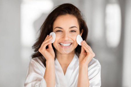 Photo for A cheerful woman playfully holds cotton pads to her cheeks, highlighting the fun in her millennial skin care routine - Royalty Free Image