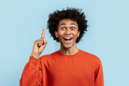 Photo for An enthusiastic young african american man in an orange sweater points upwards with a lively expression against a blue background - Royalty Free Image