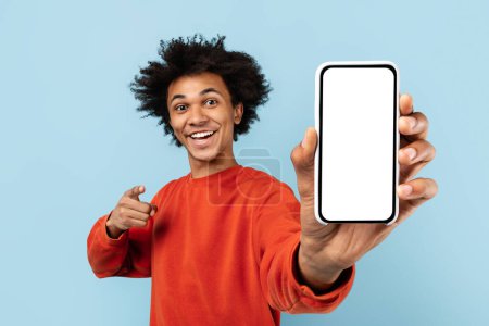 Zoomer afro-américain énergique pointant vers un écran de smartphone vierge, idéal pour la publicité d'applications, sur fond bleu isolé