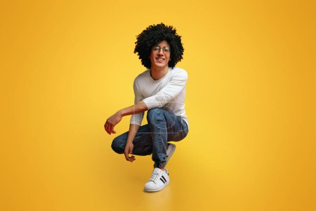 My style. Studio portrait of young black man hunkering down and smiling, orange background