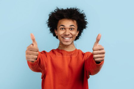 Spirited african american guy giving the thumbs up sign twice, expressing enthusiasm and approval, isolated on a blue background making a positive statement