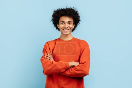 Photo for An african american young man exudes confidence and approachability with crossed arms, wearing an orange sweater, standing against a solid blue isolated background - Royalty Free Image