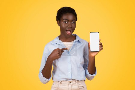 Photo for An african american woman from generation z, zoomer, points at her phones blank screen, isolated on yellow background - Royalty Free Image