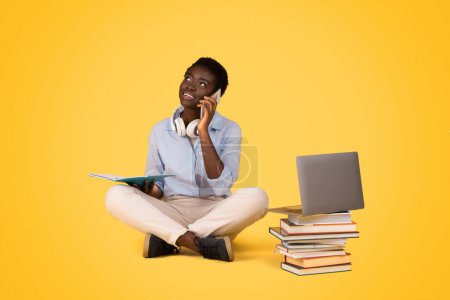 Foto de Una generación reflexiva z afroamericana se sienta con las piernas cruzadas con un libro y un ordenador portátil, contemplando, aislado en un fondo amarillo brillante, tener conversación telefónica - Imagen libre de derechos