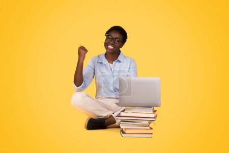Photo for Content African American woman with laptop, seated on a stack of books, showing the joy of learning and the synergy of technology and education, against a yellow background - Royalty Free Image