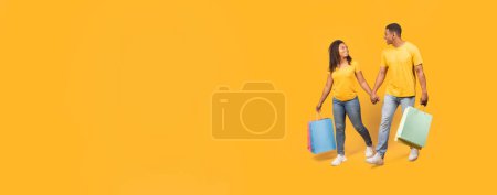 Foto de Dos personas en atuendo casual llevan coloridas bolsas de compras sobre un fondo amarillo con amplio espacio de copia, perfecto para banners web o maquetas - Imagen libre de derechos