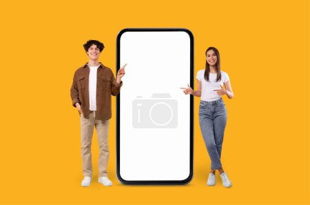 Two joyful zoomers man and woman next to an empty screen smartphone for advertising on yellow background