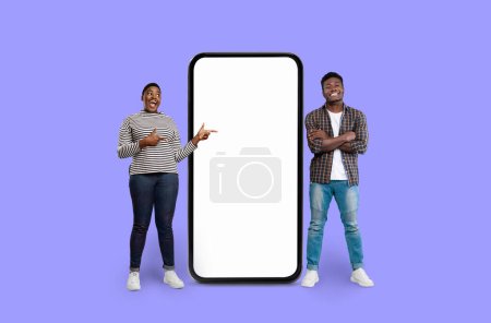 Foto de Dos personas afroamericanas haciendo gestos hacia una maqueta de teléfonos inteligentes a gran escala en violeta - Imagen libre de derechos