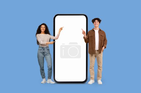 Happy young casual couple point at blank smartphone screen mockup copy space on a blue studio backdrop