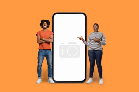 Foto de Gente afroamericana alegre que indica una gran pantalla vacía del teléfono inteligente sobre un fondo naranja vívido - Imagen libre de derechos