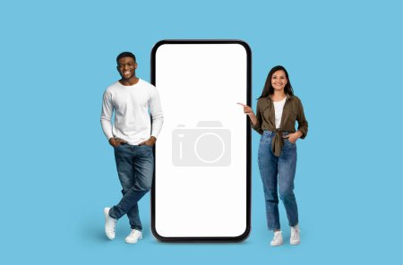 Smiling Multiracial couple pointing towards a huge mobile phone illustration on blue studio background