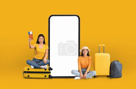 Téléchargez les photos : Deux petites amies avec des valises se tiennent à côté d'un grand smartphone, indiquant une application de voyage et de vacances ou une offre en ligne liée à la technologie numérique. Ils sont isolés sur un fond jaune - en image libre de droit