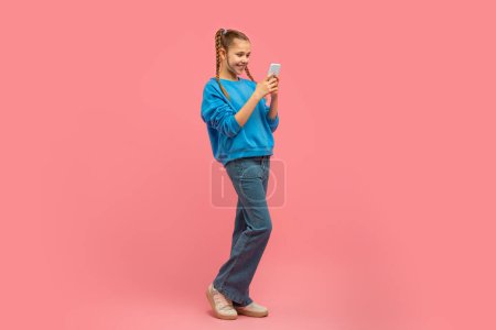 Foto de Una joven alegre con el pelo trenzado está de pie, felizmente mirando su teléfono inteligente sobre un fondo rosa - Imagen libre de derechos