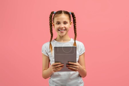 Photo for A cheerful young girl with braided hair is holding a digital tablet with both hands, standing against a vibrant pink background - Royalty Free Image
