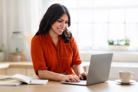 Téléchargez les photos : Heureuse femme du Moyen-Orient dans un haut orange souriant tout en tapant sur son ordinateur portable dans un cadre de cuisine lumineux - en image libre de droit