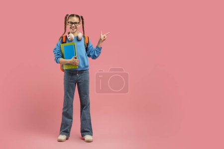 Téléchargez les photos : Joyeuse adolescente portant des lunettes portant un sac à dos et des livres pointant vers l'espace de copie sur fond rose, pleine longueur - en image libre de droit
