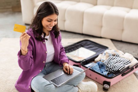 Téléchargez les photos : Une jeune femme joyeuse du Moyen-Orient assise sur le sol avec un ordinateur portable et une carte de crédit, valise ouverte à côté d'elle, représente la préparation pour le voyage - en image libre de droit