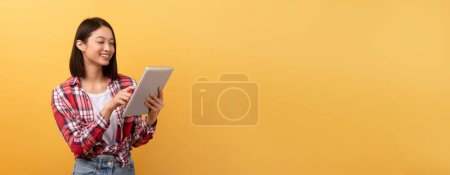 A cheerful young asian woman in plaid shirt is using a digital tablet on a vibrant yellow background with copy space