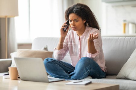 Eine schwarze Dame scheint besorgt zu sein, während sie telefoniert und auf ihren Laptop-Bildschirm blickt und ein ernsthaftes Gespräch zu Hause hervorhebt.