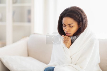 Téléchargez les photos : La femme afro-américaine est assise sur un canapé, enveloppée dans une couverture confortable. Elle semble malade, souffrant de rhume ou de grippe, toussant - en image libre de droit