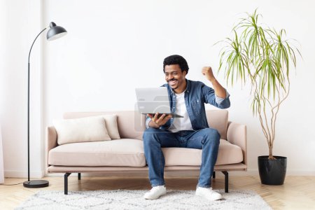 Téléchargez les photos : Homme afro-américain excité à la maison en utilisant un ordinateur portable, suggérant une expérience en ligne gagnante ou positive - en image libre de droit
