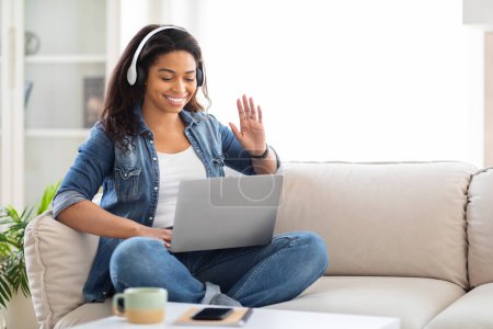 Foto de Mujer afroamericana está sentada en un sofá con una computadora portátil abierta delante de ella, escribiendo y navegando por Internet. Ella aparece enfocada y comprometida en su trabajo o actividad de ocio. - Imagen libre de derechos