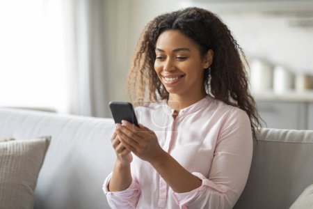 Afroamerikanerin zu Hause beschäftigt sich mit ihrem Smartphone, was einen entspannten Lebensstil und Konnektivität widerspiegelt