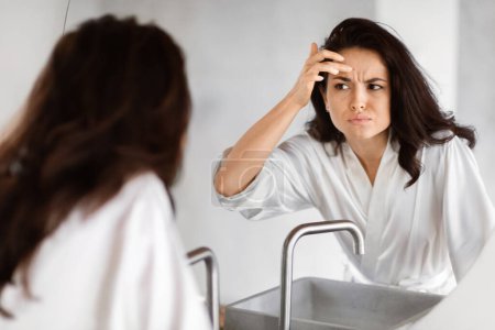 A woman in a white robe appears concerned as she examines her forehead for wrinkles in the bathroom mirror, embodying beauty anxieties