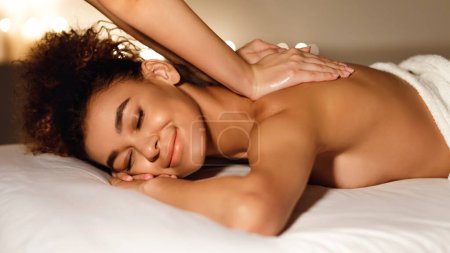 Photo for A relaxed young african american woman enjoys a soothing back massage at a spa, lying on her stomach on a massage table with a serene expression - Royalty Free Image