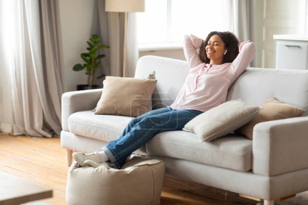 Téléchargez les photos : An african american lady enjoys a relaxing moment at home on her sofa, stretching with her eyes closed and smiling - en image libre de droit