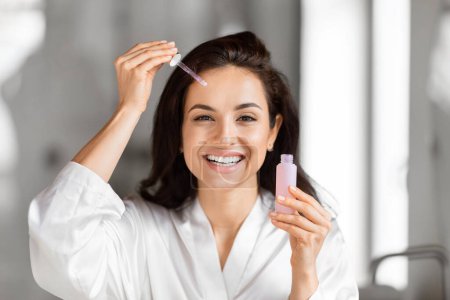 Femme souriante dans un chemisier blanc appliquant du sérum facial à partir d'une bouteille compte-gouttes sur son front, regardant la caméra, routine de soins de la peau