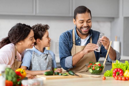 Téléchargez les photos : Un père afro-américain joyeux assaisonnant la nourriture tout en cuisinant une salade en famille, mettant en valeur les activités de cuisine et la vie domestique - en image libre de droit
