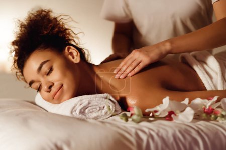 Foto de Una mujer afroamericana sonríe con placer mientras recibe un masaje en el hombro, lo que sugiere un tema de relajación dichosa - Imagen libre de derechos