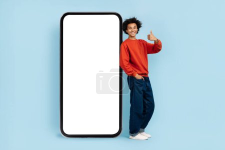 Téléchargez les photos : Un jeune homme afro-américain joyeux portant des vêtements décontractés donne un pouce vers le haut à côté d'un smartphone géant avec un écran vide - en image libre de droit