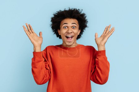 Hombre afroamericano en un suéter naranja que parece sorprendido con las manos levantadas, aislado sobre un fondo azul, retratando la sorpresa y la emoción