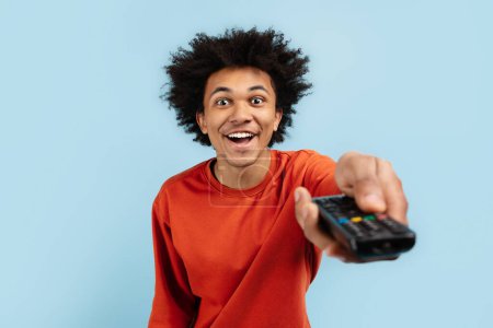 This image captures the excitement of a young black guy as he holds a TV remote control, representing leisure activities of a zoomer, isolated blue background