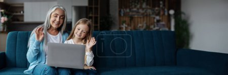 Téléchargez les photos : Petite fille européenne heureuse et femme mûre regardent l'ordinateur portable, agitant la main dans l'intérieur du salon. Rencontre à distance, appel vidéo, relation petite-fille et grand-mère à la maison, panorama avec espace de copie - en image libre de droit