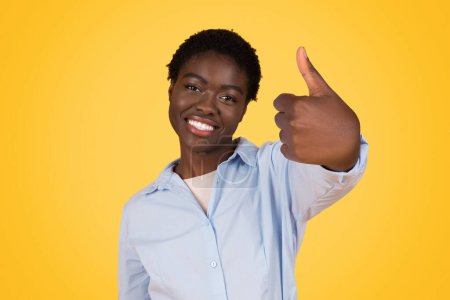 Eine lächelnde junge afrikanisch-amerikanische Frau mit kurzen Haaren gibt vor leuchtend gelbem Hintergrund ein Daumenhoch-Zeichen, das Positivität ausdrückt