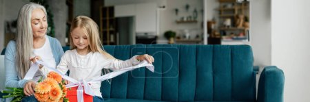 Photo for Happy caucasian small granddaughter opens box with gift from elderly grandma with bouquet of flowers on sofa in the living room. Surprise, birthday greeting and holiday celebration at home, copy space - Royalty Free Image