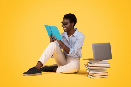 Foto de Una mujer afroamericana zoomer absorbida en la lectura sobre un fondo amarillo aislado con una computadora portátil y una pila de libros, lo que refleja un amor por el aprendizaje - Imagen libre de derechos