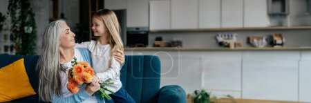 Cheerful little girl hugs european elderly lady, gives flowers and congratulates, have fun in living room interior. Birthday celebration, holiday, love, family relationship, panorama with copy space