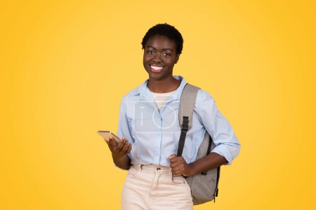 Sonriendo casualmente, una mujer afroamericana de la generación z, una zoómetra, sostiene su teléfono y su mochila, aislados en amarillo