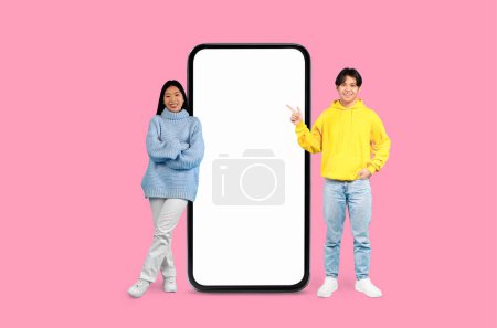 Glückliches asiatisches Paar lächelt freundlich und gestikuliert in Richtung eines leeren Smartphone-Bildschirms auf rosa Hintergrund