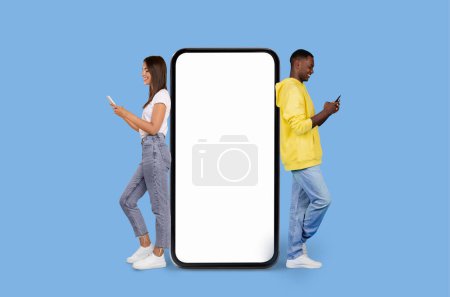 Foto de Una mujer joven y un hombre negro se paran espalda con espalda, absortos en sus teléfonos inteligentes, junto a una gran pantalla de teléfono inteligente en blanco sobre un fondo azul - Imagen libre de derechos