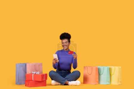 A young woman is seated cross-legged on the floor surrounded by colorful shopping bags, holding a credit card in one hand and a mobile phone in the other against a vibrant yellow backdrop