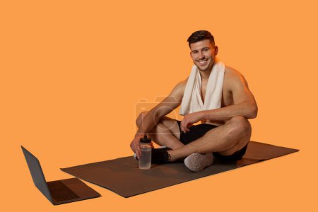 A cheerful man sits on a yoga mat with a white towel draped over his shoulders, holding a water bottle. With a laptop beside him, it appears he following an online workout or yoga session