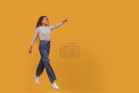 Foto de Una mujer negra alegre parece flotar mientras apunta hacia arriba, aislada sobre un fondo naranja monocromo, espacio para copiar - Imagen libre de derechos