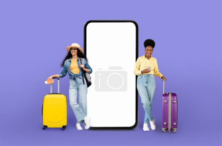 Téléchargez les photos : Deux femmes diverses avec des valises à côté d'un smartphone surdimensionné, suggérant l'utilisation d'applications de voyage, isolées sur un fond violet - en image libre de droit