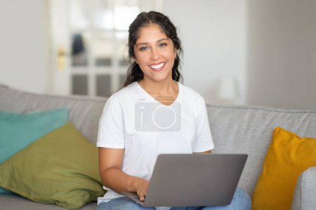 Mujer joven sonriente con freelancer portátil trabajando cómodamente desde casa, abrazando el estilo de vida laboral remoto